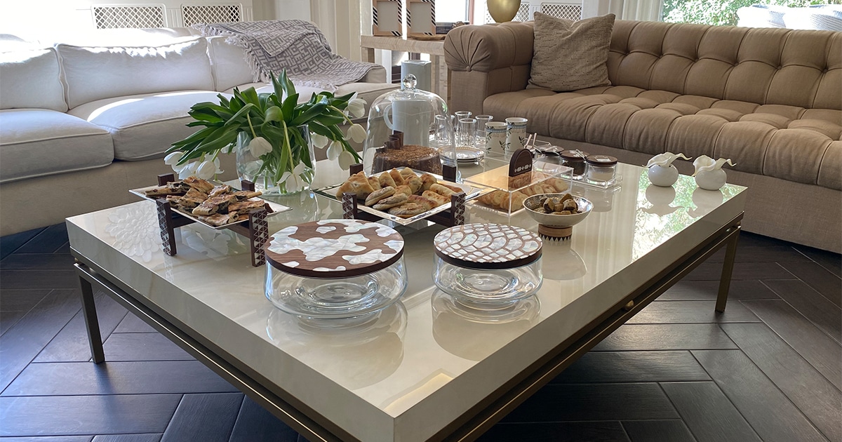 The alt text for the image you sent is: A coffee table in a living room, styled with oriental touches for a morning coffee ritual. The table is adorned with an acrylic tray with mother-of-pearl handles, an olive mug, matching dessert plates, a porcelain thermos, a tiny vase with a white flower, and aluminum plates with mother-of-pearl designs elevated on wooden risers.